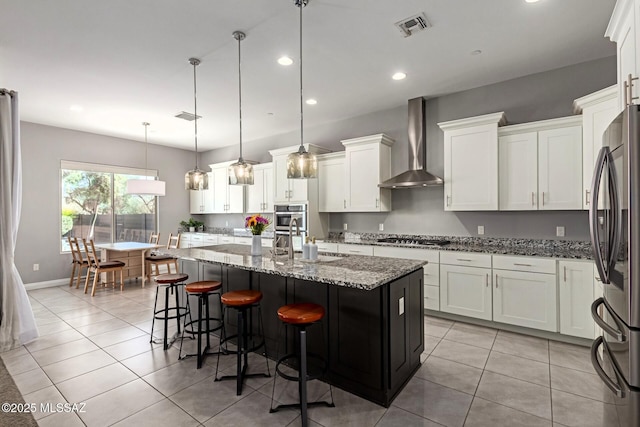 kitchen featuring pendant lighting, appliances with stainless steel finishes, white cabinetry, an island with sink, and wall chimney exhaust hood