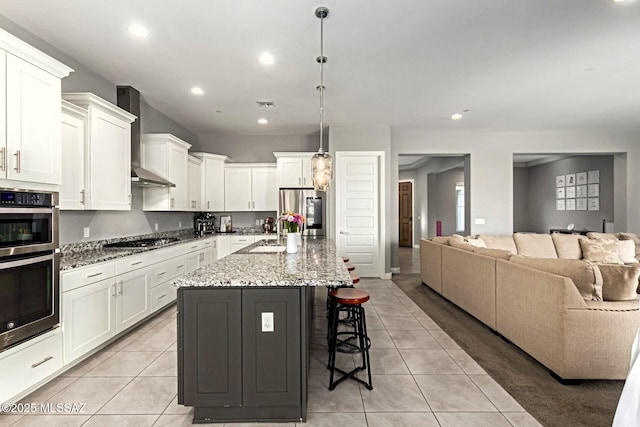 kitchen featuring hanging light fixtures, appliances with stainless steel finishes, wall chimney range hood, a kitchen island with sink, and white cabinets