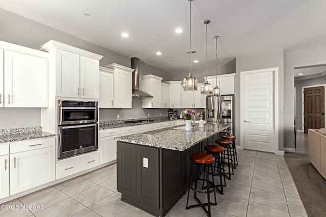 kitchen featuring hanging light fixtures, an island with sink, stone counters, wall chimney range hood, and white cabinets