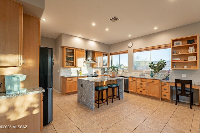 kitchen featuring wall chimney range hood, black refrigerator, a kitchen breakfast bar, a center island, and light stone countertops