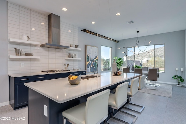 kitchen featuring a breakfast bar, sink, wall chimney range hood, hanging light fixtures, and an island with sink