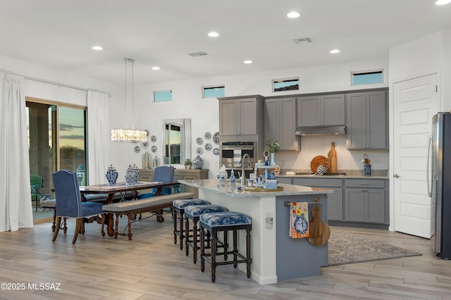 kitchen with decorative light fixtures, an island with sink, light stone counters, stainless steel appliances, and light wood-type flooring