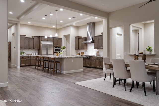 kitchen featuring wall chimney exhaust hood, stainless steel built in fridge, an island with sink, and backsplash