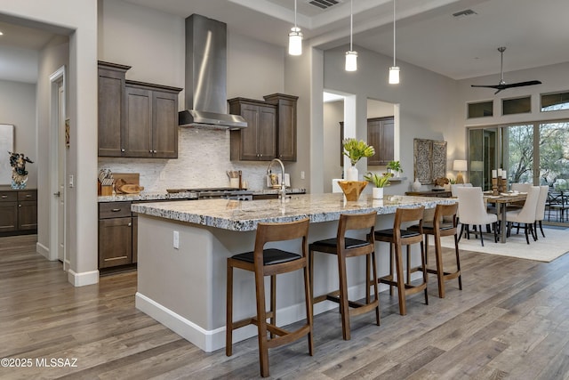 kitchen with a kitchen bar, hanging light fixtures, wall chimney range hood, light stone counters, and a center island with sink