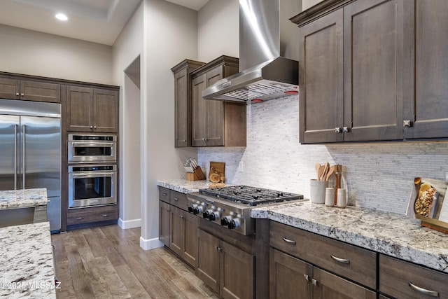 kitchen with wall chimney exhaust hood, light stone counters, dark brown cabinets, light hardwood / wood-style flooring, and appliances with stainless steel finishes