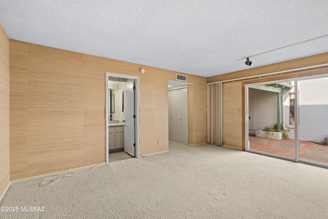 unfurnished bedroom featuring a textured ceiling, wooden walls, a closet, light colored carpet, and ensuite bathroom