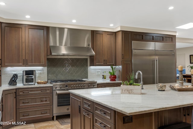 kitchen featuring light stone countertops, premium appliances, wall chimney range hood, and tasteful backsplash