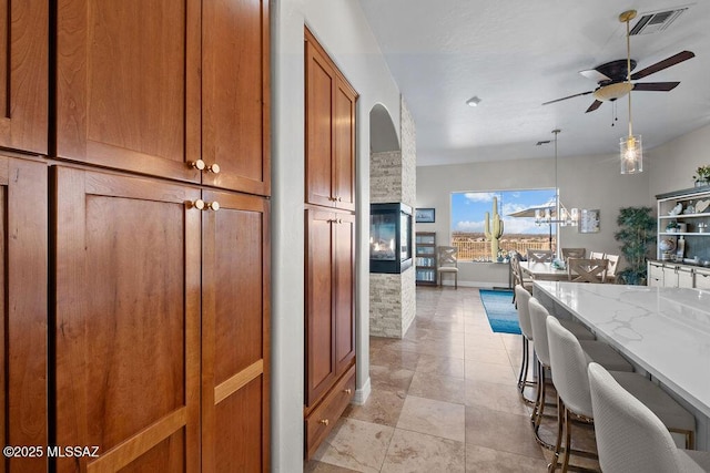 kitchen featuring a stone fireplace, light stone counters, a breakfast bar, visible vents, and brown cabinetry