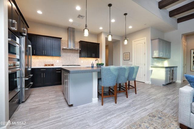 kitchen featuring decorative backsplash, beamed ceiling, appliances with stainless steel finishes, a breakfast bar area, and wall chimney exhaust hood