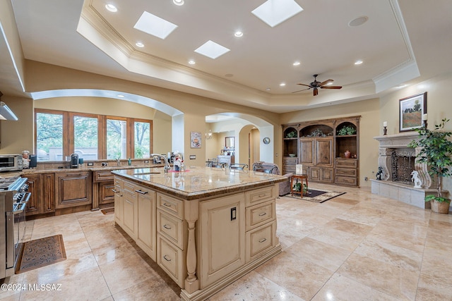 kitchen featuring an island with sink, a tiled fireplace, a tray ceiling, and light stone countertops