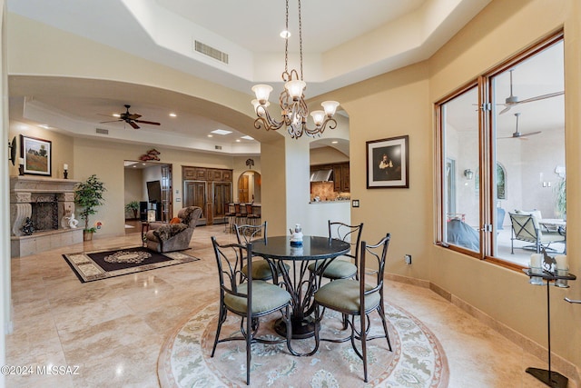 dining space with a tiled fireplace, a raised ceiling, and ceiling fan with notable chandelier