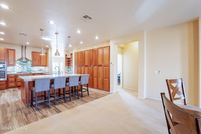 kitchen featuring pendant lighting, wall chimney range hood, a breakfast bar area, an island with sink, and decorative backsplash