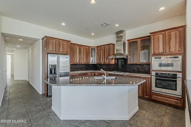 kitchen with dark stone counters, a kitchen island with sink, stainless steel appliances, a sink, and wall chimney range hood