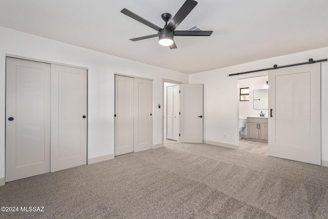 unfurnished bedroom with two closets, ceiling fan, light colored carpet, ensuite bathroom, and a barn door