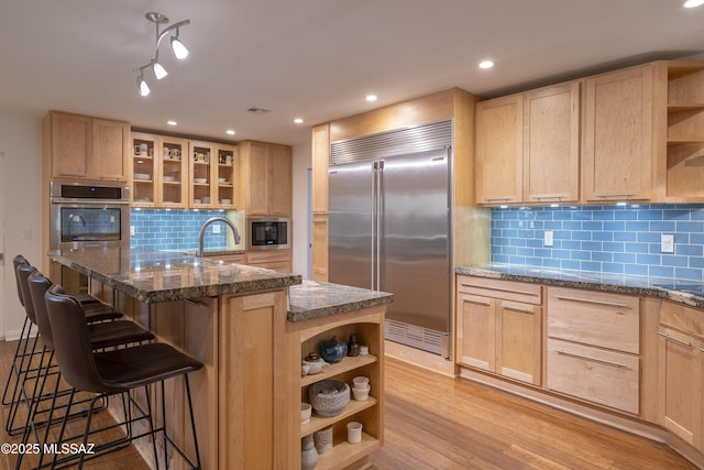 kitchen featuring a kitchen breakfast bar, a kitchen island with sink, built in appliances, and light brown cabinetry