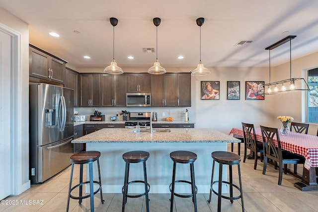 kitchen featuring decorative light fixtures, a kitchen island with sink, light stone counters, stainless steel appliances, and dark brown cabinets