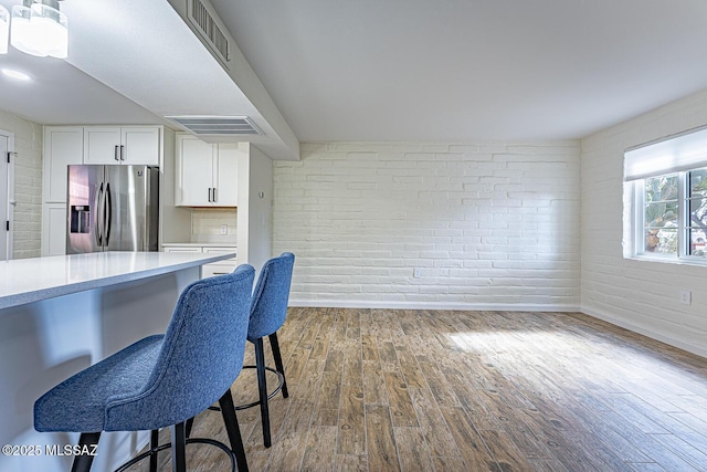 kitchen featuring stainless steel refrigerator with ice dispenser, brick wall, wood-type flooring, white cabinets, and a breakfast bar