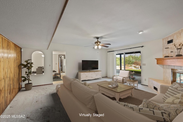 carpeted living room with ceiling fan, wooden walls, a textured ceiling, and a tiled fireplace