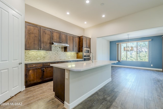 kitchen with stainless steel microwave, light hardwood / wood-style floors, black electric stovetop, hanging light fixtures, and a kitchen island with sink