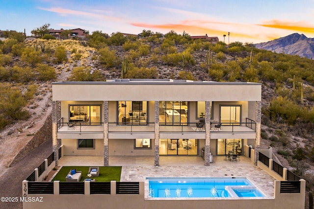 back house at dusk featuring a mountain view, a swimming pool with hot tub, a balcony, and a patio