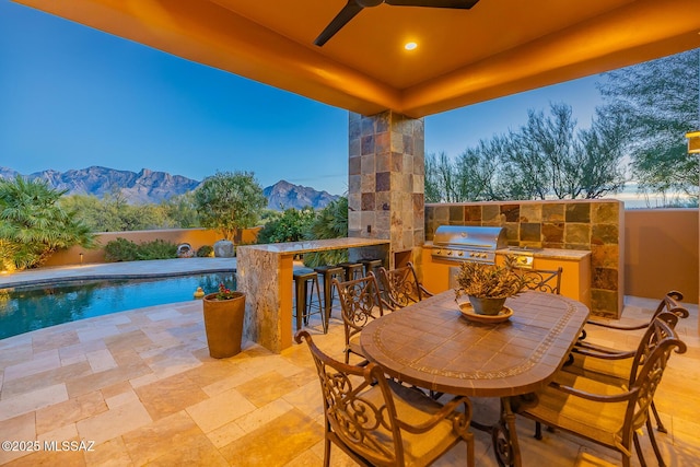 patio terrace at dusk featuring a grill, an outdoor bar, ceiling fan, a fenced in pool, and a mountain view