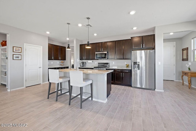 kitchen featuring pendant lighting, dark brown cabinets, stainless steel appliances, an island with sink, and a kitchen bar