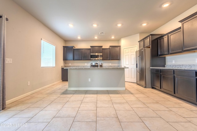 kitchen featuring dark brown cabinets, light tile patterned floors, appliances with stainless steel finishes, light stone countertops, and a kitchen island with sink