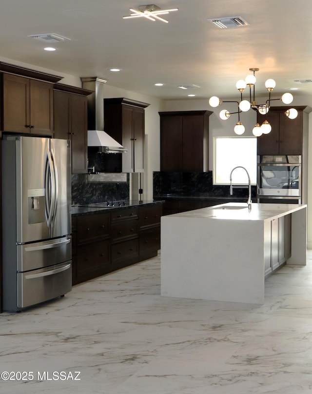 kitchen with appliances with stainless steel finishes, pendant lighting, sink, a center island with sink, and wall chimney exhaust hood