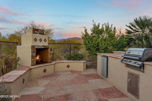 patio terrace at dusk with grilling area, a mountain view, area for grilling, and an outdoor stone fireplace