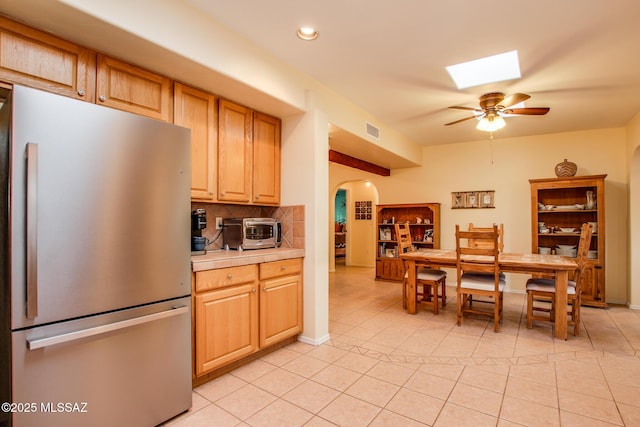 kitchen with light tile patterned flooring, a skylight, tasteful backsplash, stainless steel fridge, and light brown cabinets