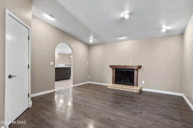 unfurnished living room featuring arched walkways, a fireplace with raised hearth, wood finished floors, and baseboards