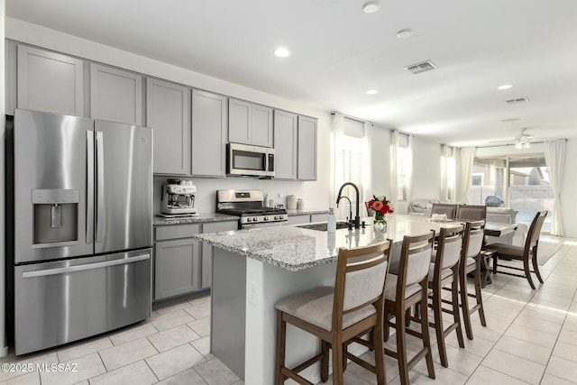 kitchen featuring light tile patterned floors, gray cabinets, appliances with stainless steel finishes, light stone countertops, and a center island with sink