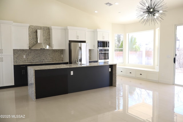 kitchen featuring stainless steel appliances, visible vents, white cabinets, wall chimney range hood, and an island with sink