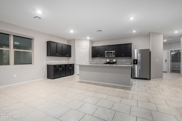 kitchen with light tile patterned floors, sink, stainless steel appliances, light stone countertops, and a center island with sink