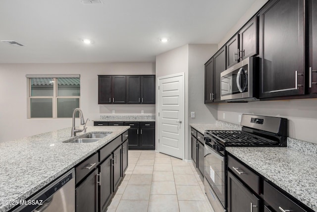 kitchen with appliances with stainless steel finishes, sink, light tile patterned floors, dark brown cabinetry, and light stone countertops