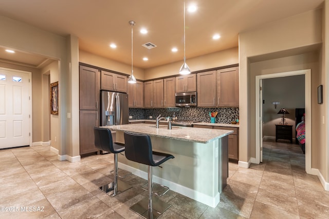 kitchen featuring appliances with stainless steel finishes, hanging light fixtures, backsplash, light stone countertops, and a center island with sink