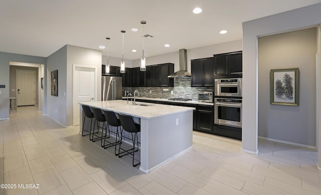 kitchen with wall chimney exhaust hood, sink, light stone counters, appliances with stainless steel finishes, and an island with sink