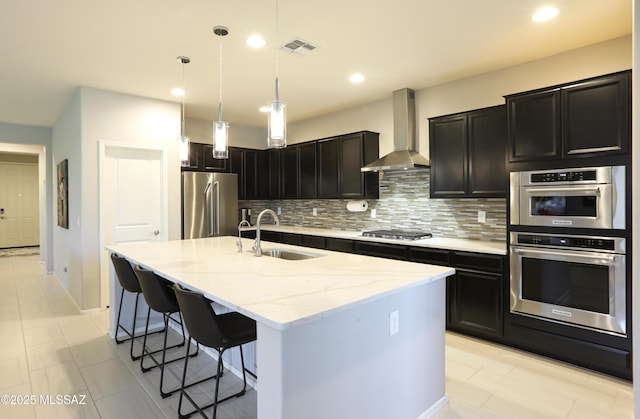 kitchen with sink, wall chimney range hood, hanging light fixtures, stainless steel appliances, and a center island with sink