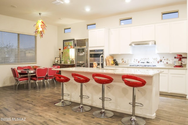 kitchen with white cabinets, appliances with stainless steel finishes, hanging light fixtures, light wood-type flooring, and a center island with sink