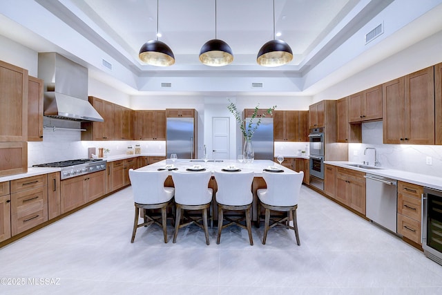 kitchen with appliances with stainless steel finishes, wall chimney range hood, decorative light fixtures, an island with sink, and a breakfast bar area