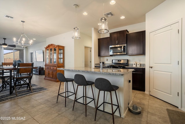 kitchen featuring hanging light fixtures, stainless steel appliances, dark brown cabinetry, light stone countertops, and a center island with sink