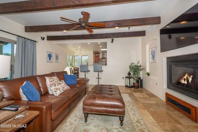 living area with a glass covered fireplace, beam ceiling, ceiling fan, and stone finish floor