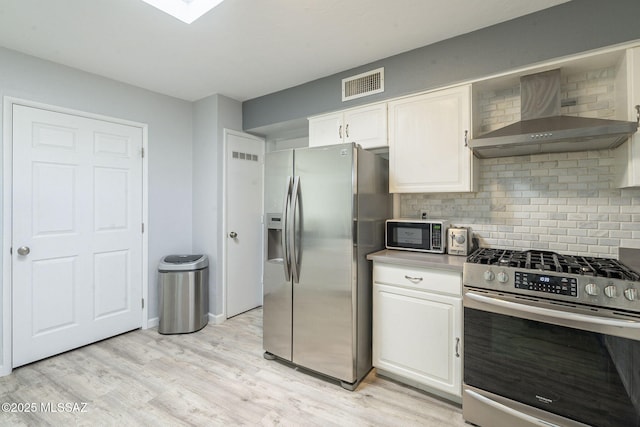 kitchen with light wood-type flooring, appliances with stainless steel finishes, white cabinets, wall chimney range hood, and backsplash