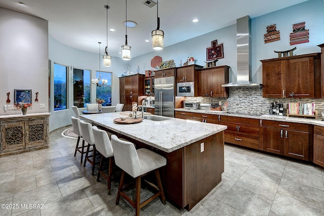 kitchen with sink, a kitchen island with sink, built in appliances, decorative light fixtures, and wall chimney exhaust hood