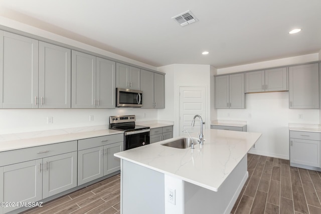 kitchen with sink, gray cabinets, appliances with stainless steel finishes, light stone countertops, and an island with sink