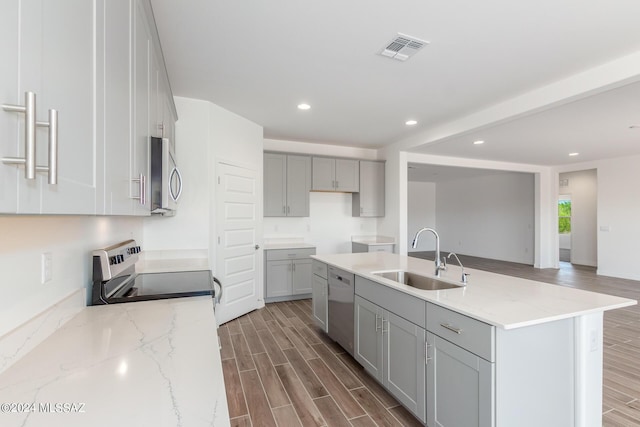 kitchen featuring an island with sink, sink, gray cabinetry, stainless steel appliances, and light stone countertops