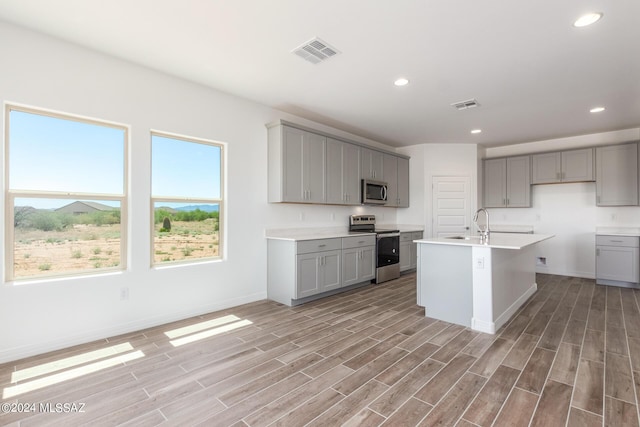 kitchen featuring appliances with stainless steel finishes, gray cabinetry, a center island with sink, and light wood-type flooring