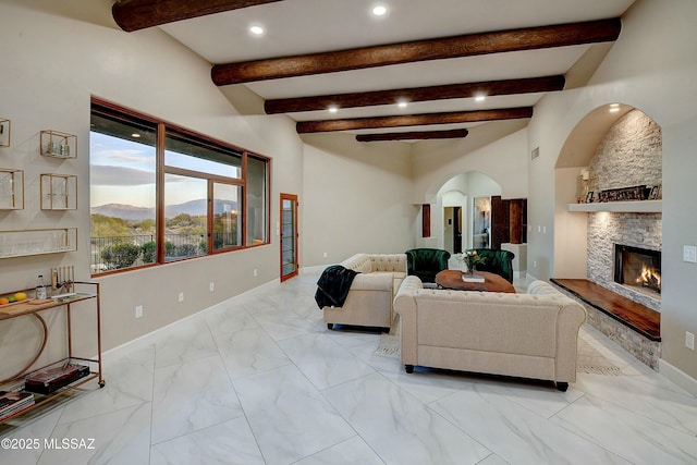 living area with baseboards, arched walkways, marble finish floor, a stone fireplace, and beam ceiling