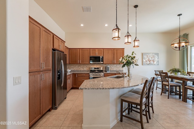 kitchen with a kitchen bar, light stone counters, hanging light fixtures, a center island with sink, and appliances with stainless steel finishes