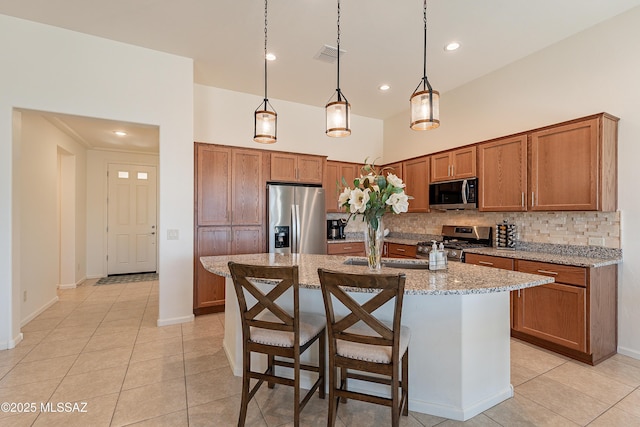 kitchen with hanging light fixtures, light stone countertops, appliances with stainless steel finishes, and a kitchen island with sink